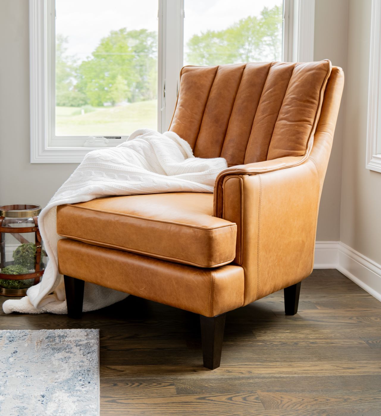 The Vanderbilt Chair in brown leather sits in a corner with a blanket draped across it near a rug from Penny Mustard