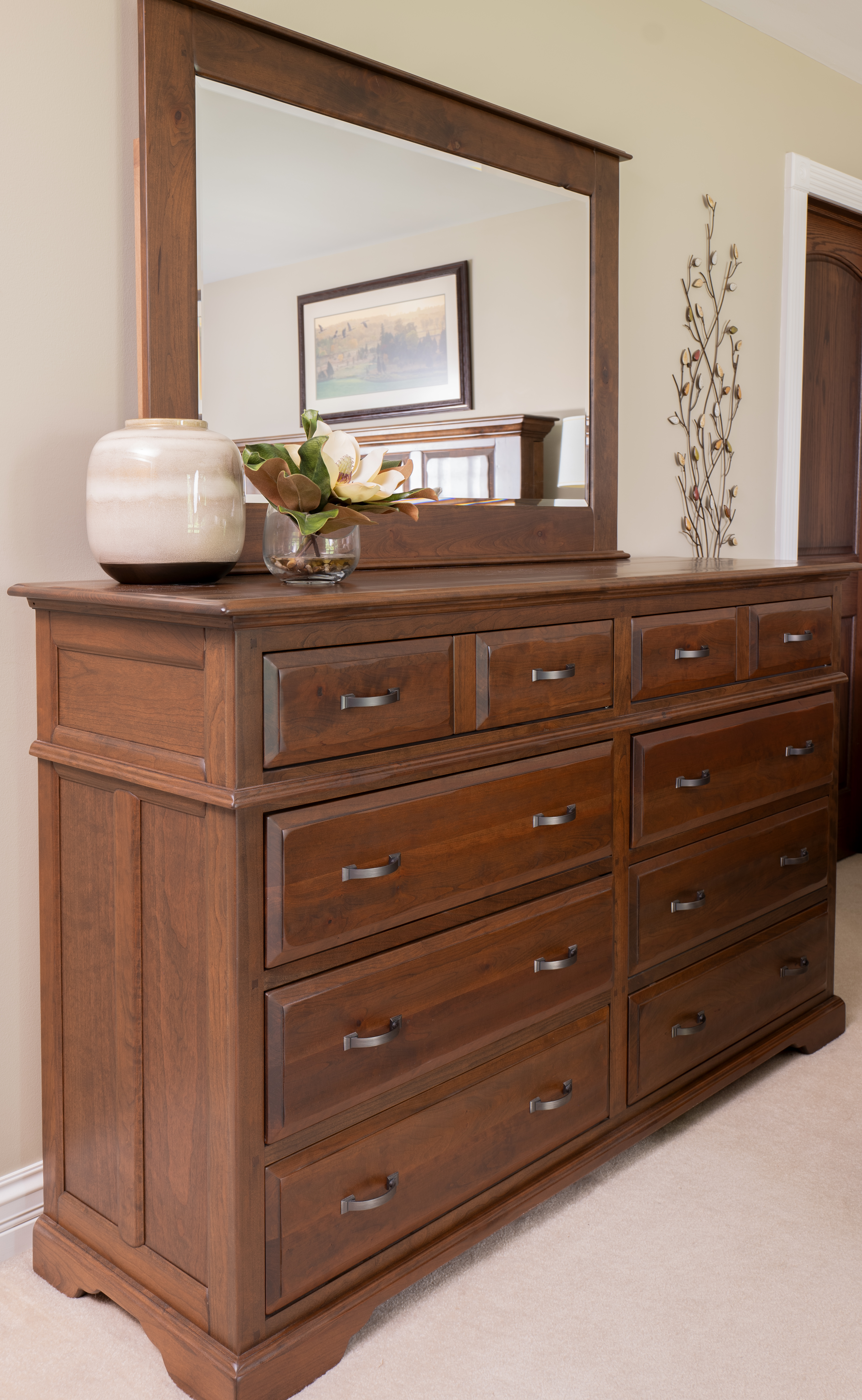 A large multi-drawer chest of drawers in a medium brown with a large mirror sits in a master bedroom.