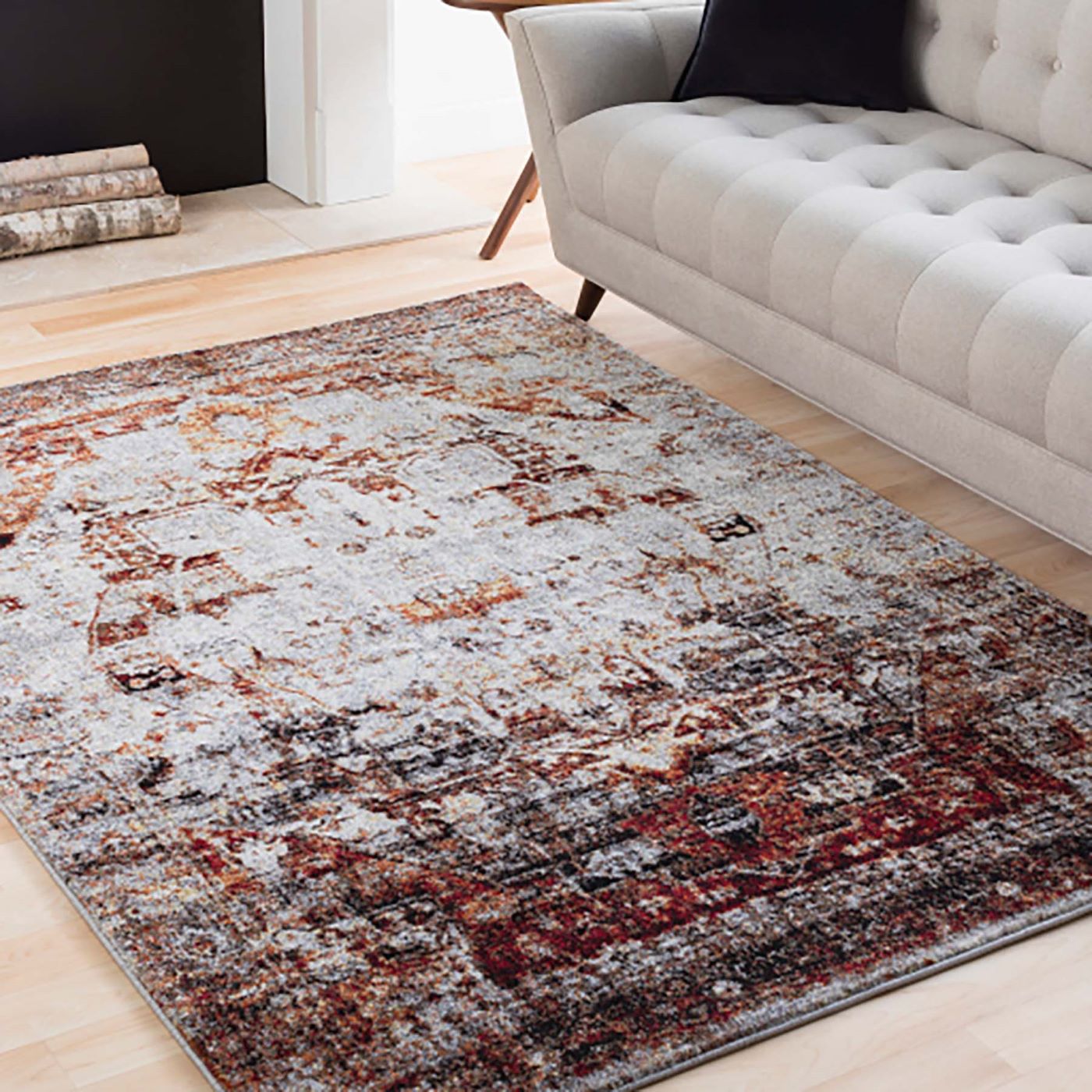 The Serapi Rug in grey, rust, and black sits on a light brown wooden floor next to a grey upholstered sofa