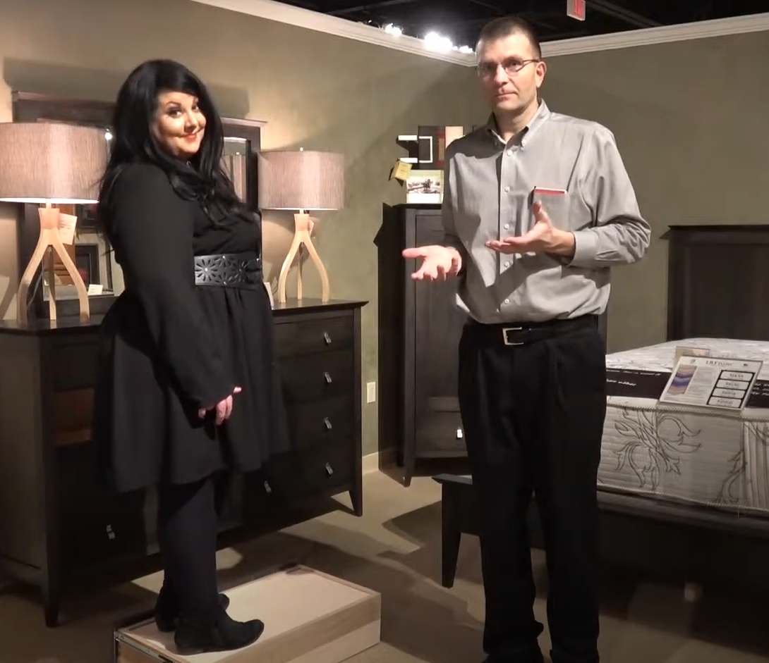 Nicole Titschler and one of the Huth brothers stand together inside a showcased bedroom setup in a Penny Mustard showroom