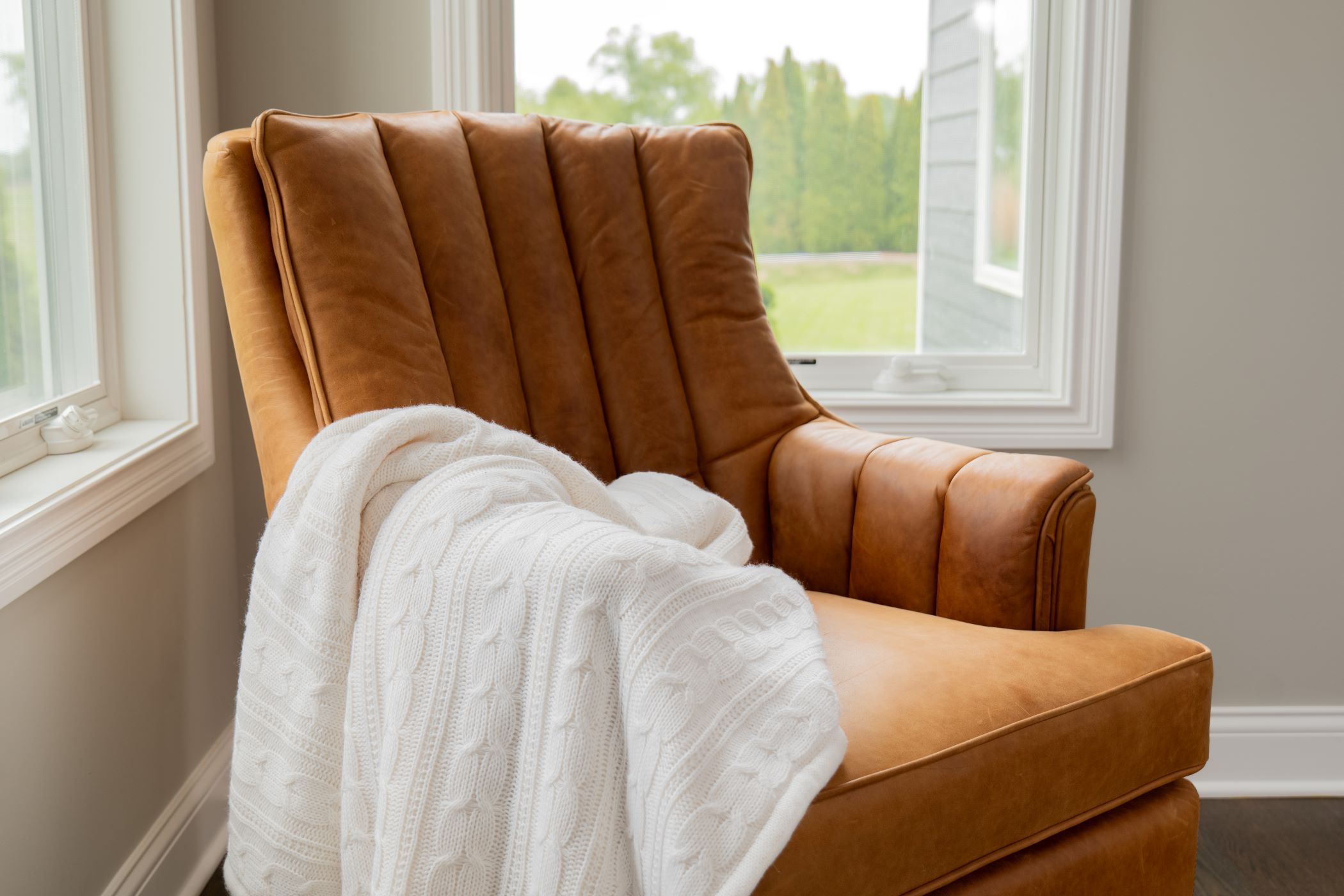 The Vanderbilt Chair in light brown leather sits in the corner between two windows with a blanket draped over the arm